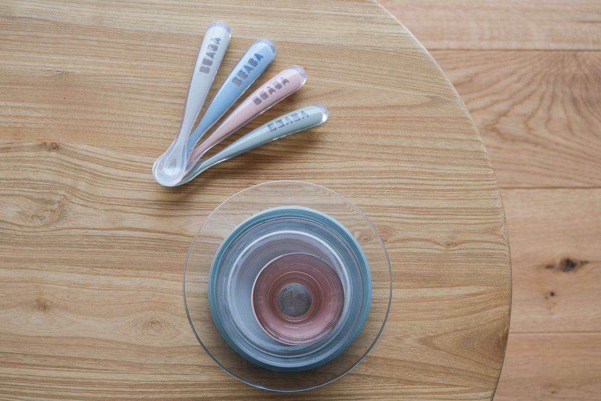 Cztery pastelowe łyżeczki Béaba obok miseczek na okrągłym drewnianym stoliku, widok z góry.